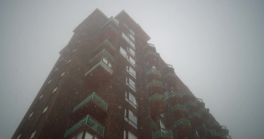 Snow falls around a tall apartment or hotel under a cloudy sky