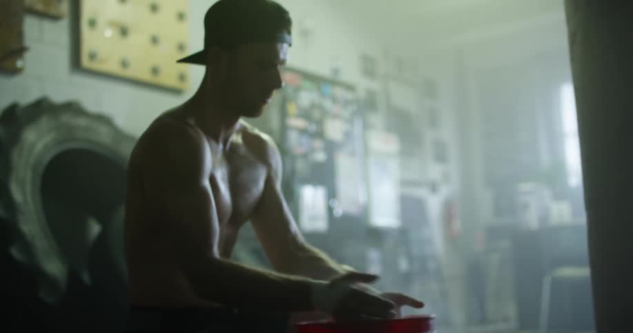 A shirtless man in a gym kneels to reach into a bucket and powder his hands