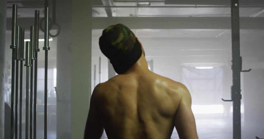 Young shirtless muscular man walks towards fitness equipment in a dimly lit gym