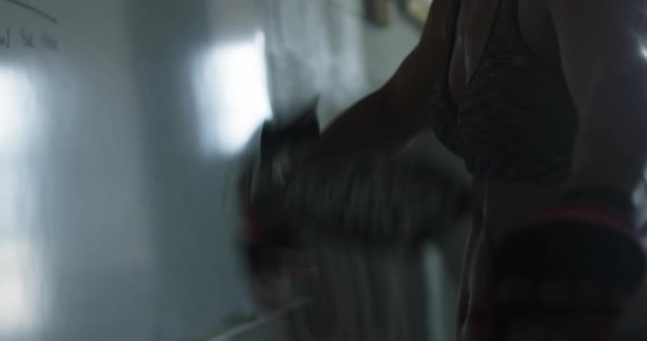 A sweaty woman in athletic wear and gloves unwinds her wristwrap as she stands in a dimly lit room