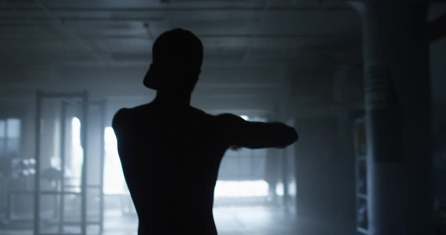 A shirtless young man does stretches in a dimly lit gym as a light flashes repeatedly