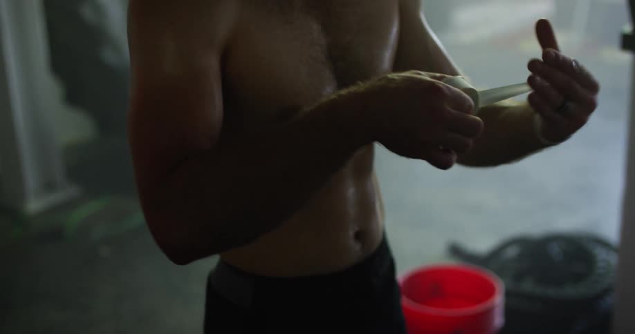 A shirtless man wraps his hand in boxing hand wraps as he stands in a gym