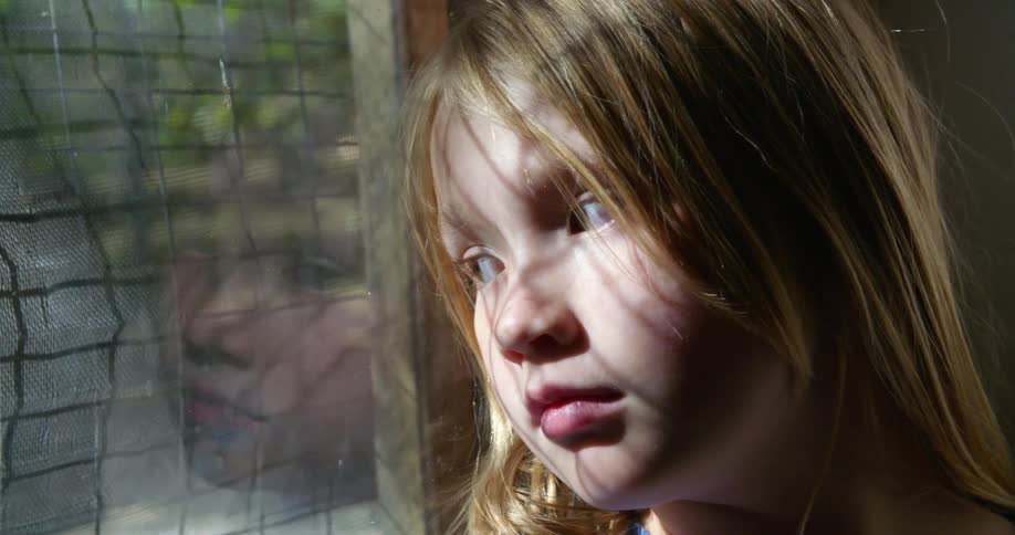A small child leaning on a window looking sad