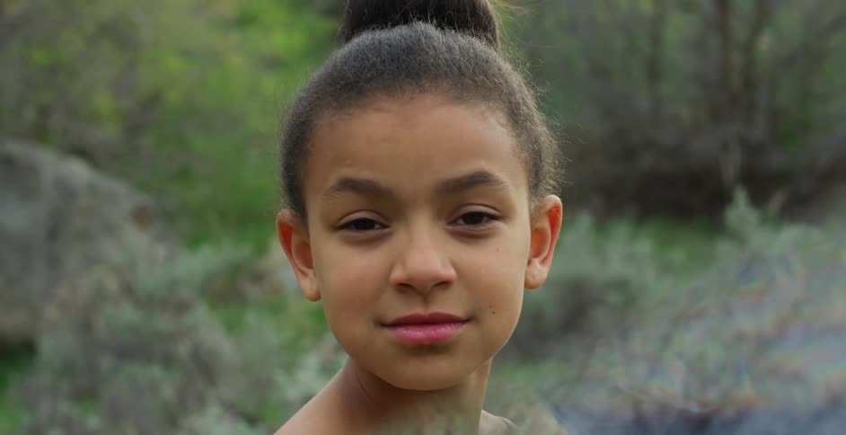 A young black girl stands outdoors and looks on with a subtle smile on her face