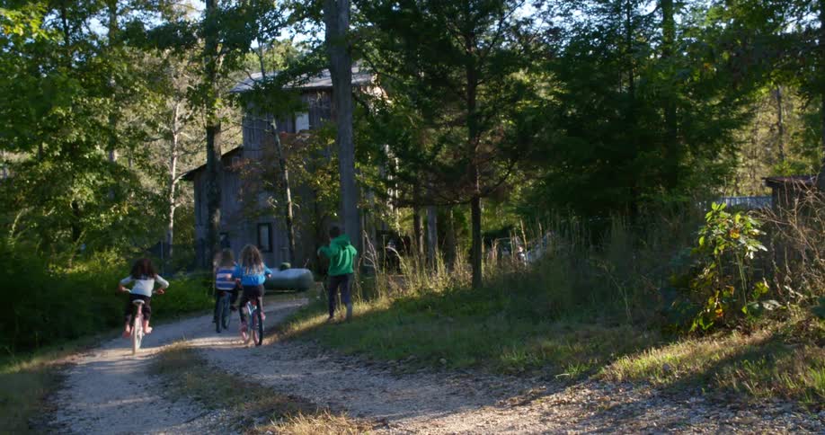 Three female girls jumps on their bicycles and ride towards the house on a dirt driveway as the boy runs after them in a chase to the finish