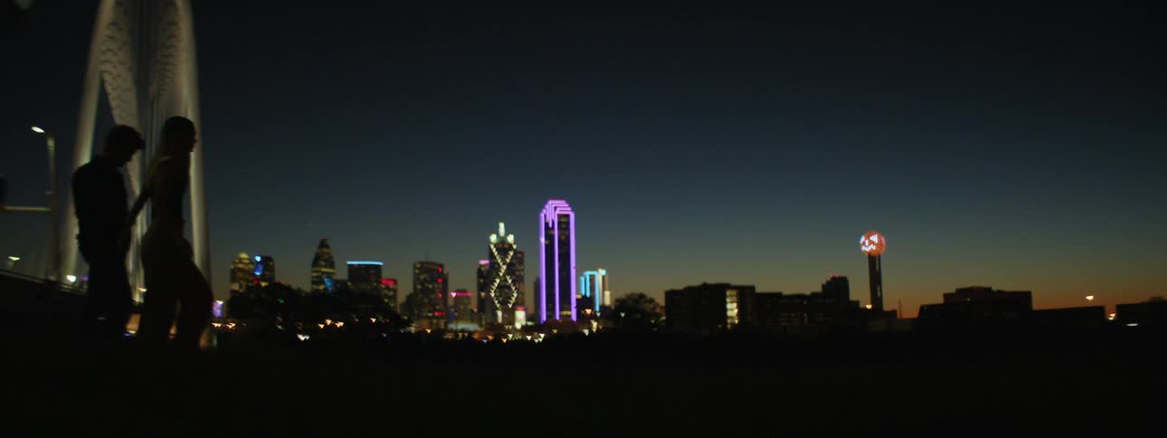 Man and woman holding hands and walking are silhouetted against the night sky and city skyline