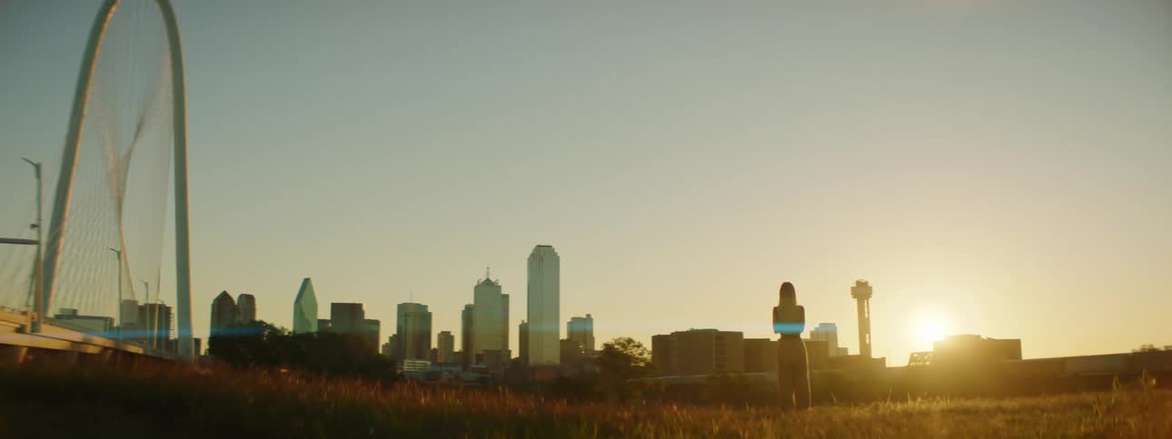 A woman looks towards distant skyscrapers as she stands in a field outside of a city during the day