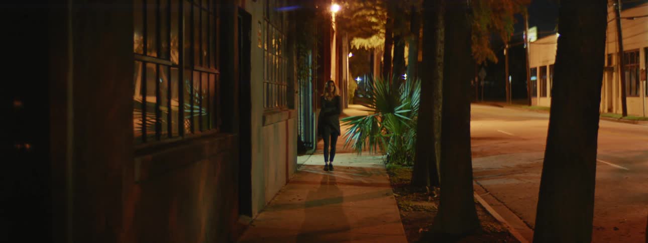 A woman holds her outer shirt closed as she walks down a city sidewalk at night