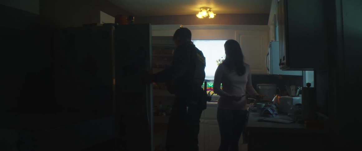 A man in a border patrol uniform enters a kitchen and checks a refrigerator before turning to argue with a woman cooking on the stove