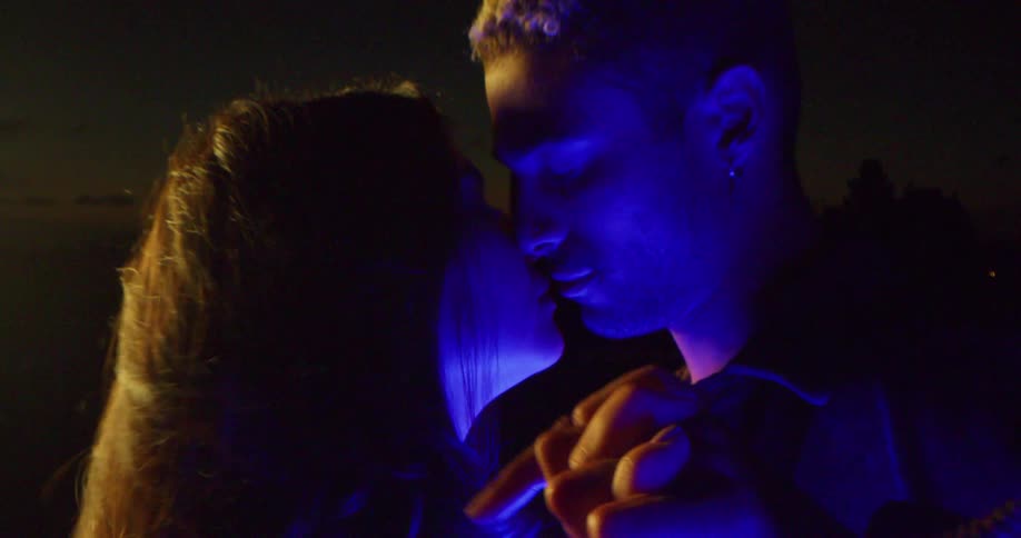 Couple lit by blue light stand face to face holding hands with skyline barely seen on horizon at night