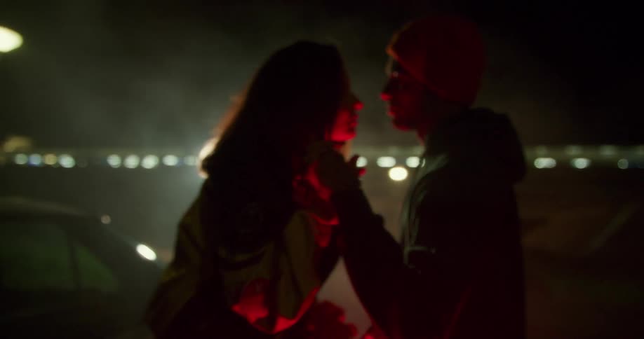 A man and woman are illuminated by the red tail lights of a vehicle as they look into each others eyes and hold hands outside the vehicle at night