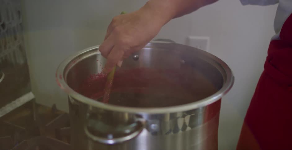 Person stirs red steaming red sauce in large pot on stove top with wooden utensil