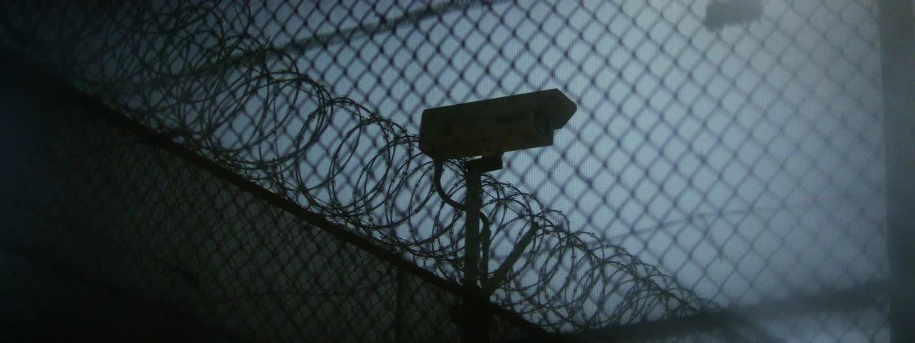 A security camera mounted on a fence with coiled barb wire on the top of it