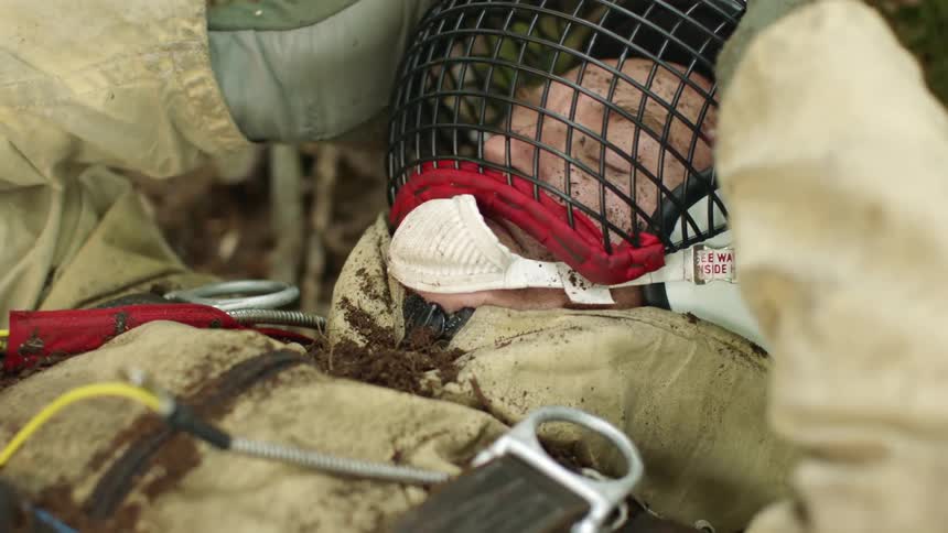 A man in a harness and personal protective equipment lies on the ground while covered in dirt before slowly sitting up
