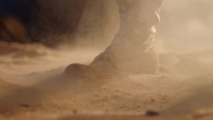 A persons foot wearing boots standing on sand and dust