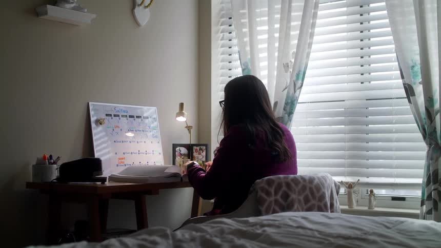 A woman sits at a bedroom desk turning the pages of a book
