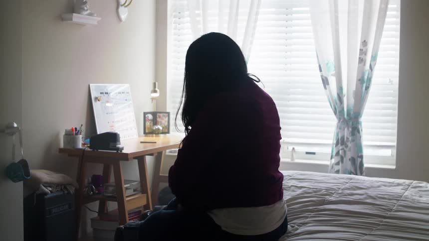 A young woman puts on her outer shirt as she sits on a bed in a bedroom