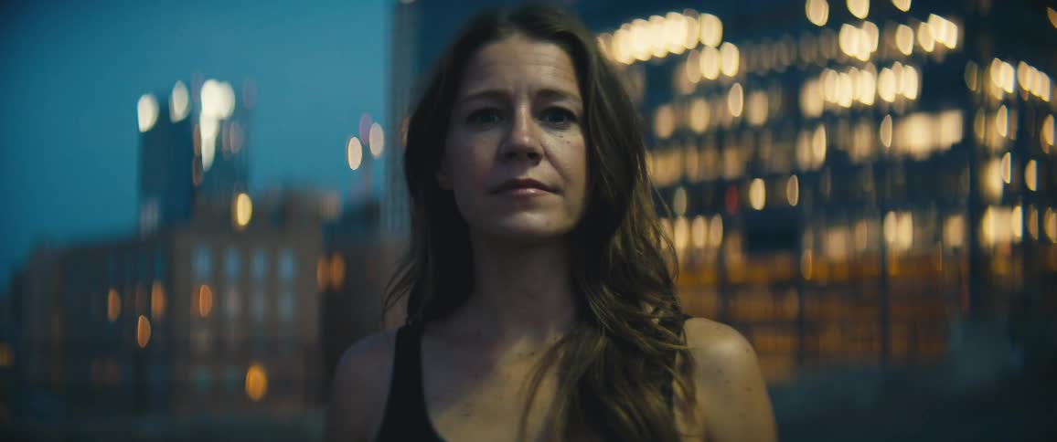 Middle aged woman with brown hair faces the camera with eye contact in front of an illuminated high rise building