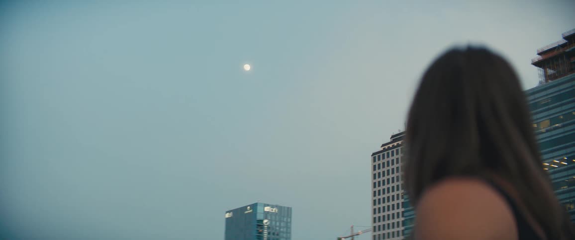 A woman looks up at the moon in the distance as day turns to night in a city
