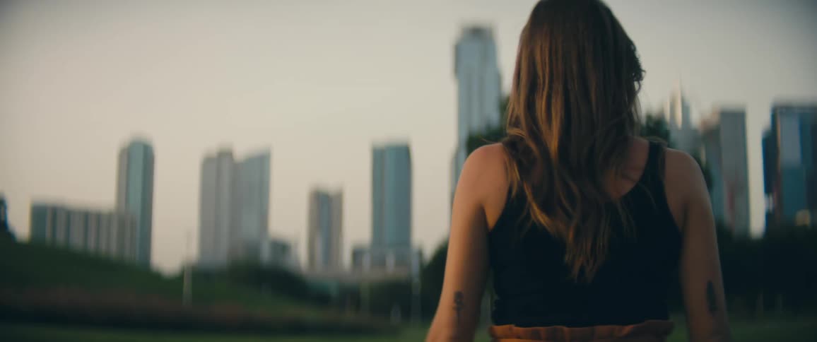 Woman with brown hair walks on green grass in public park with city skyline in the background