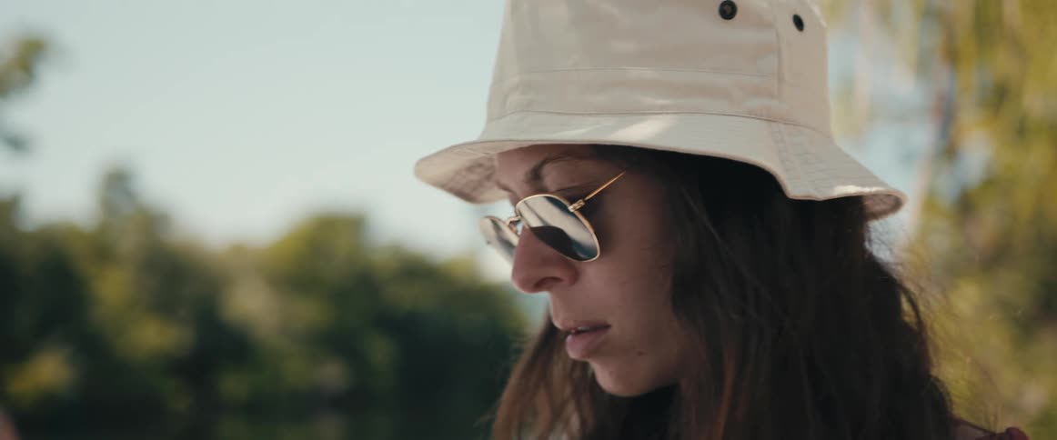 Woman stands in shades and hat near water viewing mobile phone