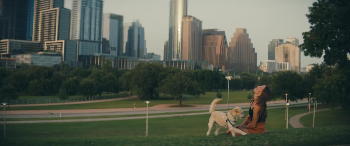 Woman with brown shorts sitting on green grass pets a white dog in public park with tall buildings in the background