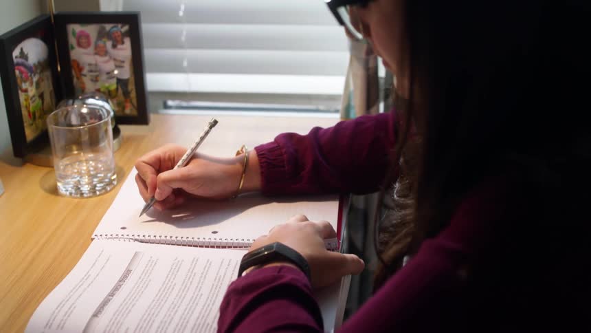 Girl with a sweater and glasses sits at a desk and writes on a notebook