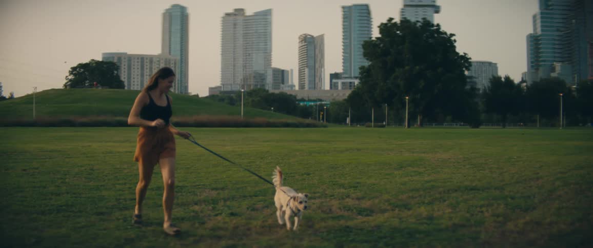 Woman with brown hair and shorts runs on green grass with a white dog in public park with city skyline in the background
