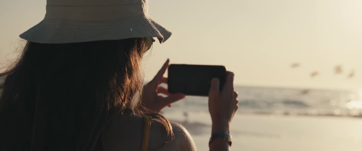 Woman with a hat and and glasses takes pictures of sea with seagulls flying over the beach