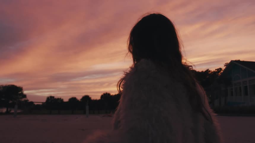 A white female in a furry jacket walks along a beach at sunset