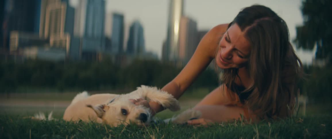 A woman scratches the stomach of her white terrier dog as they lie on the grass in a park near downtown