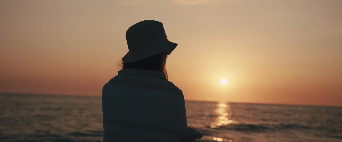 A female in a hat stands at the edge of the water looking at a sunset