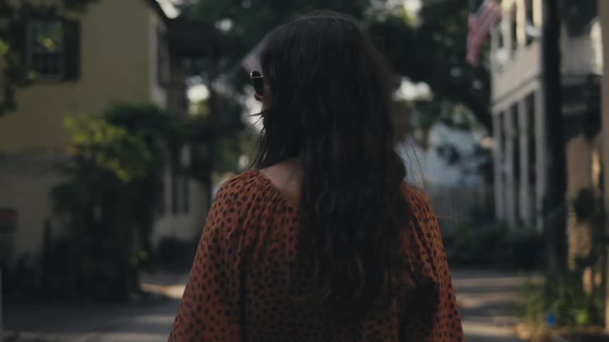 A woman in sunglasses walks down a walkway between houses during the day