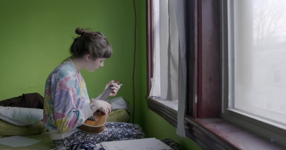 A woman is playing a mini guitar by the window