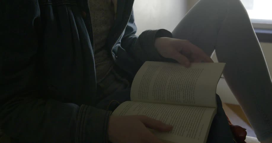 A woman sits next to a window reading a book