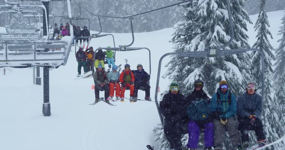 Skiers and snowboarders sit four to a chair on a ski lift as snow falls down
