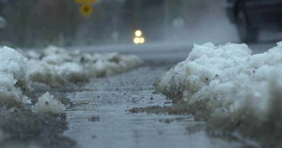 A black vehicle slowly drives down a slick road as rain falls and melts snow on the shoulder