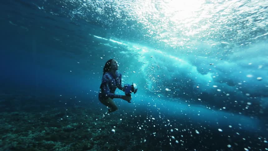 A woman holds a camera underwater with waves overhead