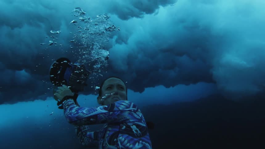 A woman swims along a reef with a camera as heavy waves roll overhead