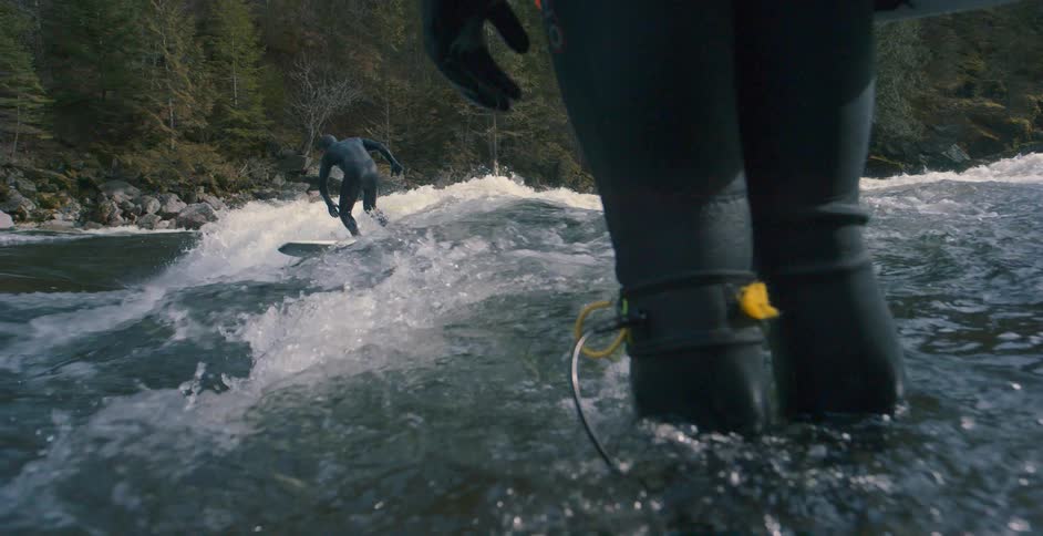 A man standing in a river watches as another man stands on the rapids on a surf board