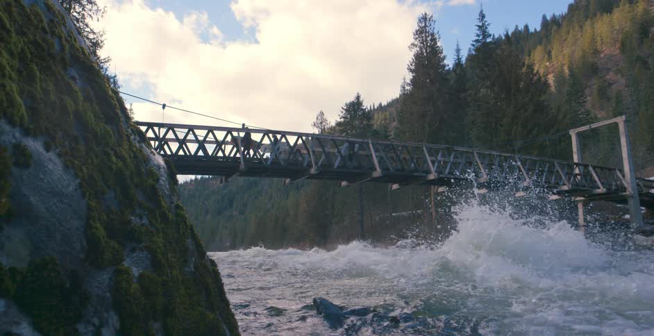Two people walk on a bridge that spans a rushing river