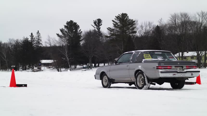 A older car slowly pulls up to a line between two traffic cones in a snow covered field