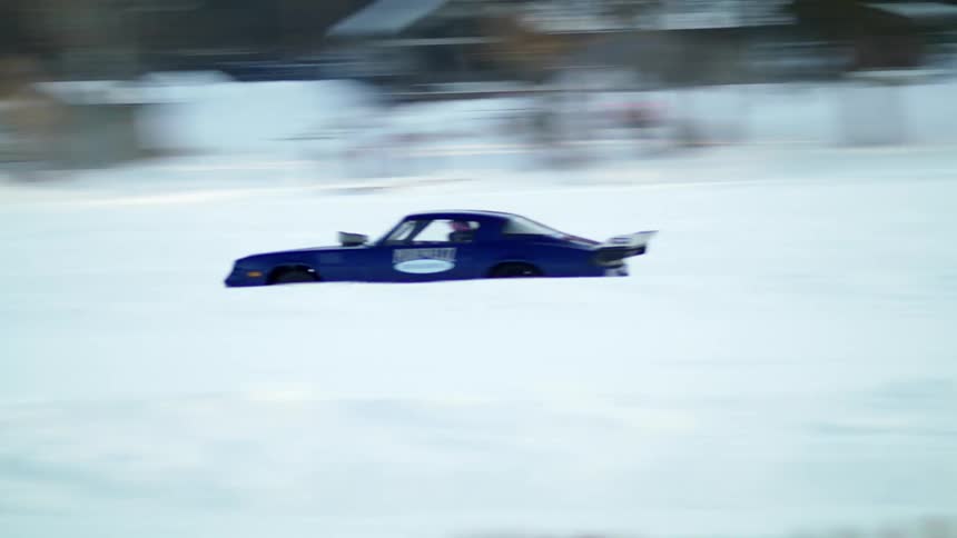 A pair of cars drag race across a field covered in snow as spectators stand by their own vehicles and watch