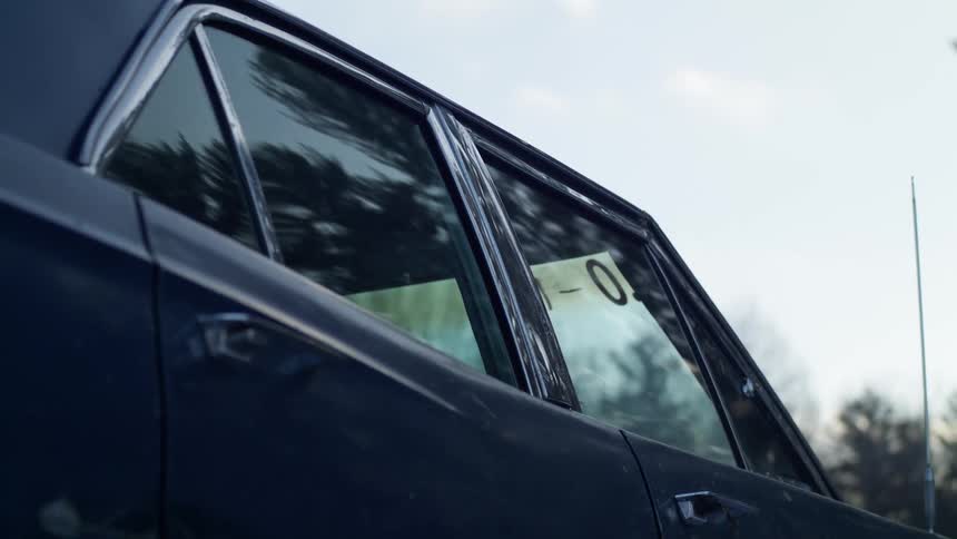 An older dark blue sedan is parked outside near a line a trees