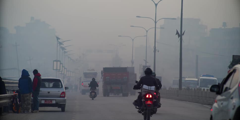Cars and motorcycles drive down a city street and two people stand at the side of the road