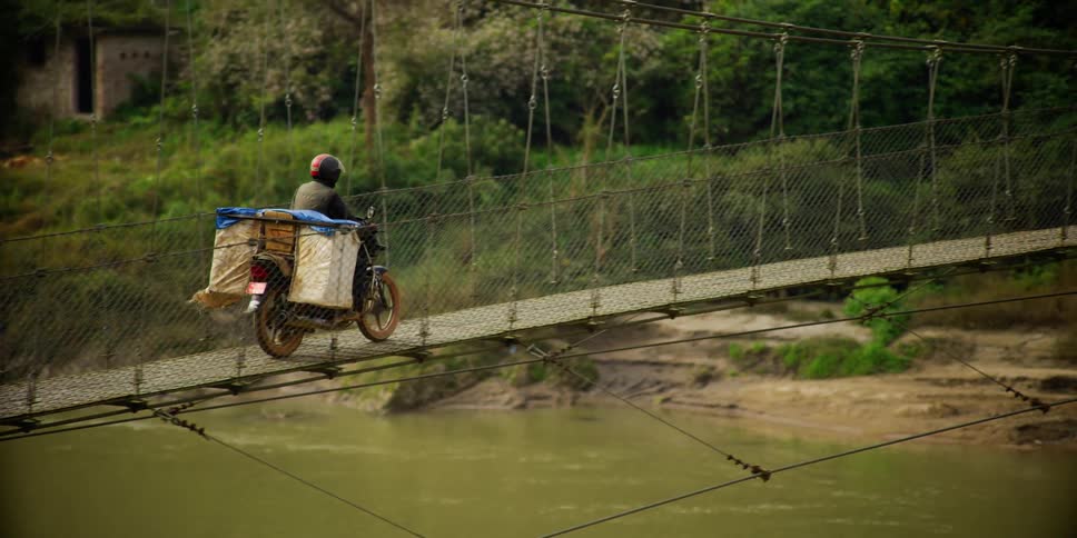 A person in a village driving a motorcycle over a very narrow suspension bridge