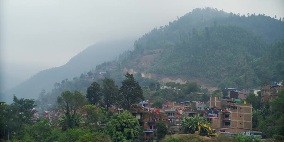 A village with buildings surrounded by plants and trees with a hill and the sky in the background