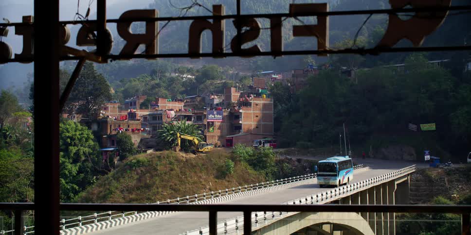 A large blue bus drives on a bridge that runs past an apartment building in a developing village