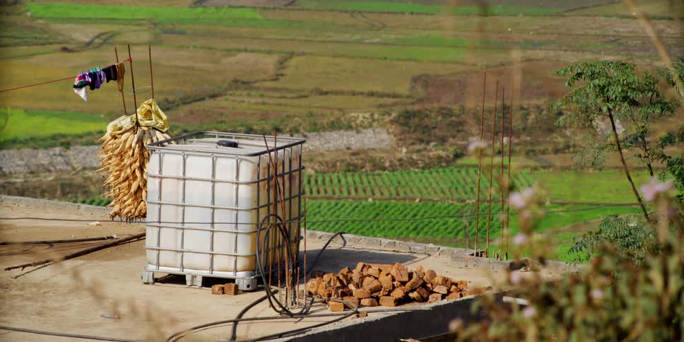 A portable water tank next to a pile of bricks on top of a roof outlooking a farm field
