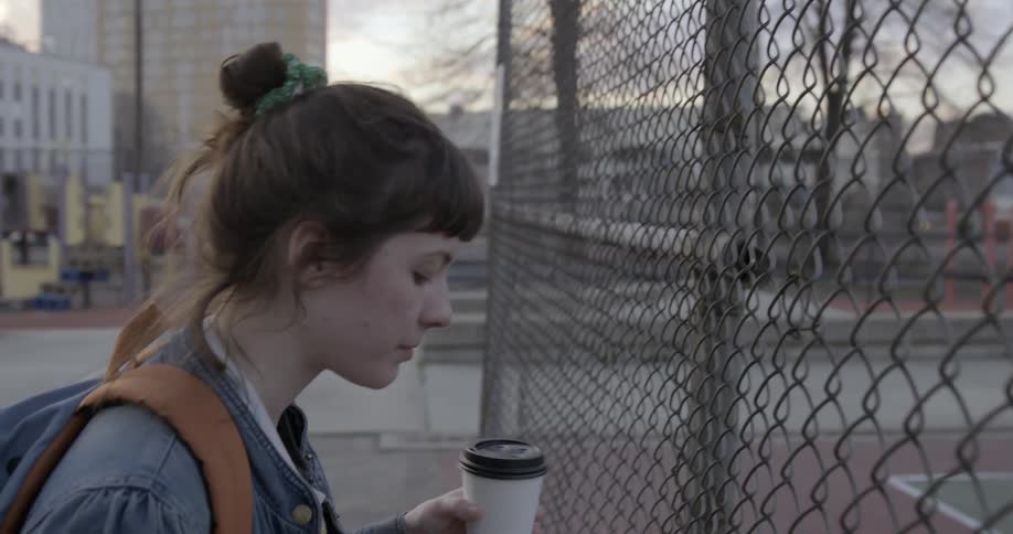 A woman stands by a fence drinking from a cup